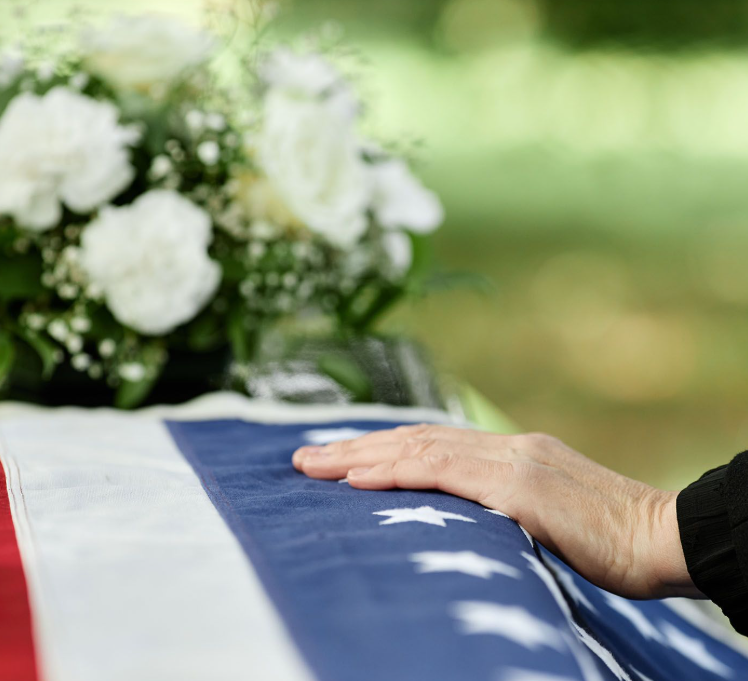 A hand resting on an American flag-draped casket beside white flowers
